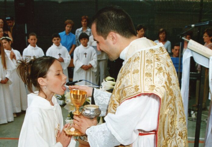 A young girl in a white alb receives First Holy Communion from a smiling priest